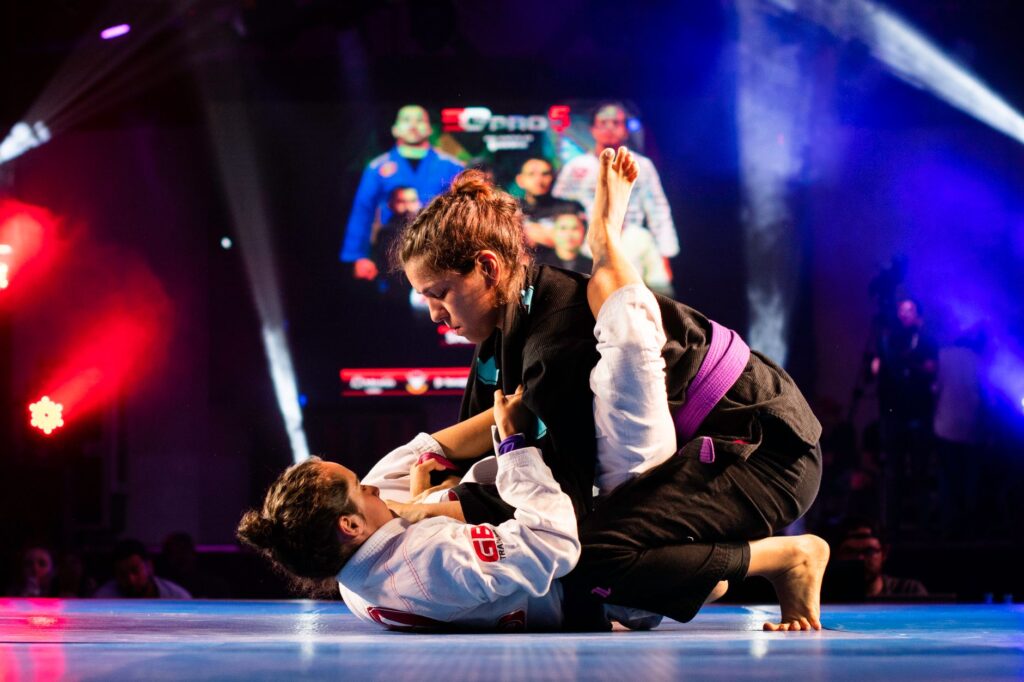 Two athletes compete in a gi jiu-jitsu match on a lit competition stage