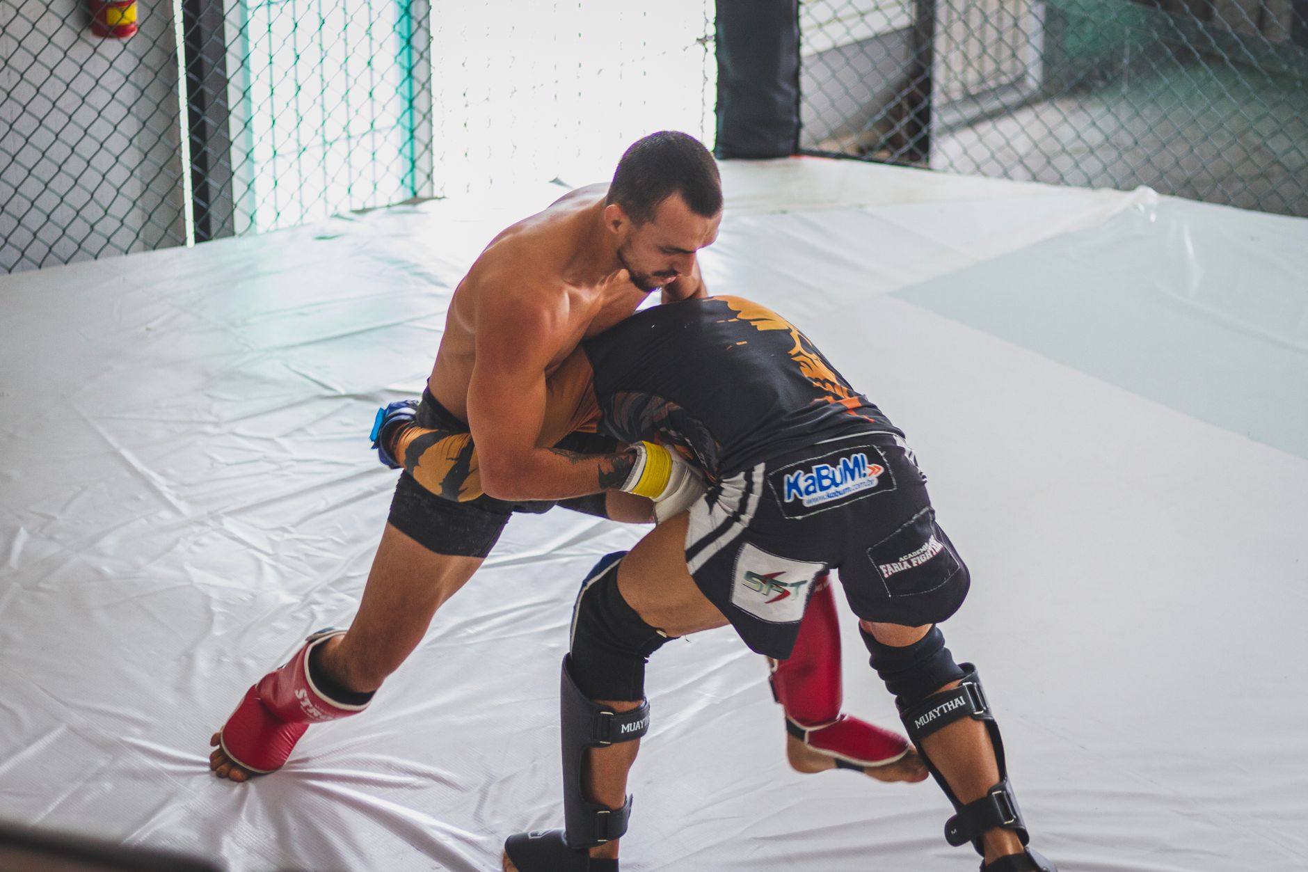 Two fighters clinching in MMA sparring with shin guards and gloves in a cage