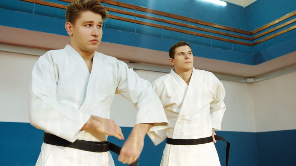 Two judoka wearing white gis and black belts standing in a dojo